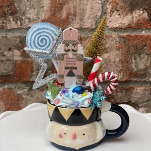 May include: A whimsical Christmas-themed mug featuring a nutcracker design. The mug is filled with whipped cream, candy, and a miniature Christmas tree. A blue and white swirled lollipop and a silver glitter star add to the festive decor.