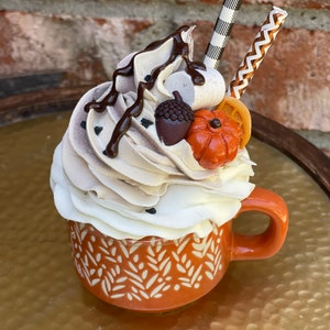 May include: An orange ceramic mug with a white leaf pattern, filled with a decorative whipped cream topping, chocolate drizzle, and fall-themed accents. The mug features a handle and sits on a gold surface. The topping includes a small orange pumpkin, an acorn, and two decorative straws.