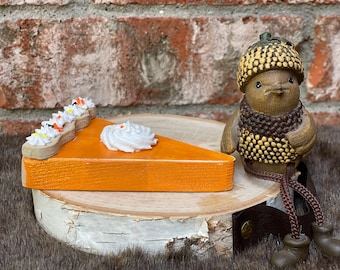 May include: A wooden slice of orange pumpkin pie topped with white whipped cream and a small acorn-shaped figurine wearing a textured cap and brown boots. The pie features a tan crust edge with white dollops and orange sprinkles.