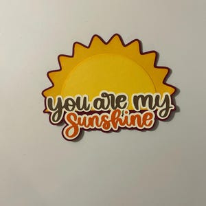 May include: A layered papercraft sun with a yellow center and a dark orange outline. The sun's rays are a darker orange. The words "You are my Sunshine" are layered on the sun in brown and orange script.