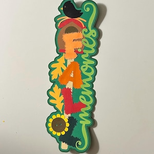 May include: A vertical layered paper cutout sign featuring the text "FALL memories" in textured orange, red, and dark green letters. The design includes a black bird, a brown birdhouse, yellow oak leaves, and a sunflower with a brown button center.