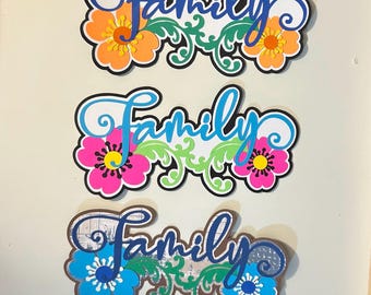 May include: Three layered wall signs each featuring the word Family in cursive script. The top sign has orange flowers, the middle has pink flowers, and the bottom has blue flowers. Each sign includes green leafy scrollwork and a black outline.