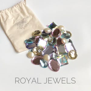 May include: A small white drawstring bag with the text "FizzyBoo" printed on it, filled with iridescent and pearlescent gems. The gems are in various shapes and sizes, including round, oval, and square. The bag is sitting on a white surface. The text "ROYAL JEWELS" is printed below the bag.