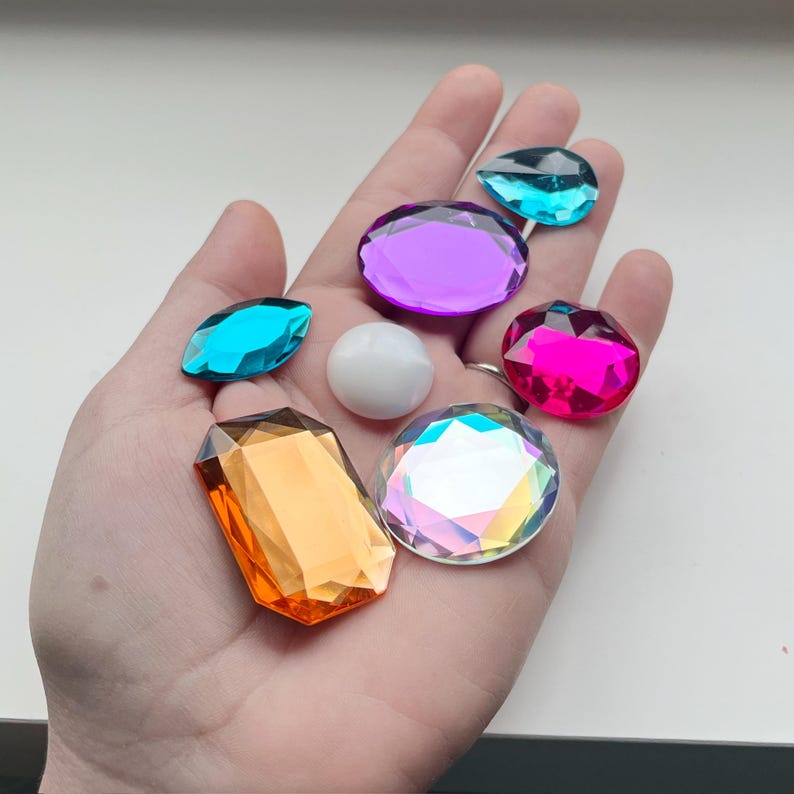 Op de afbeelding: Een hand die een verscheidenheid aan gekleurde edelstenen vasthoudt, waaronder een paarse, roze, blauwe, oranje en iriserende edelsteen. Er is ook een witte parel-achtige edelsteen.