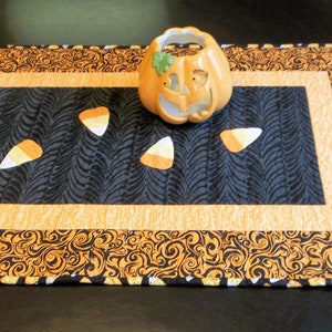 Può includere: Un runner da tavolo di Halloween nero e arancione con un pannello centrale nero con un motivo a spirale e un bordo in tessuto arancione e nero. Cinque decorazioni di mais dolce arancione e bianco sono sparse sul runner, e al centro c'è un portacandele a forma di zucca in ceramica.