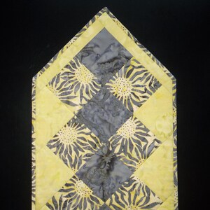 May include: A yellow and gray quilted table runner with a sunflower pattern. The runner is made of fabric squares sewn together in a diamond pattern.