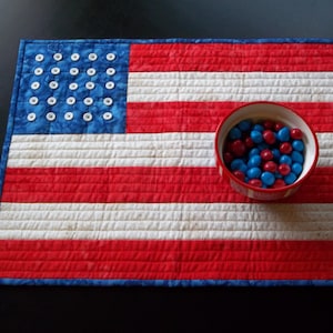 May include: A red, white, and blue American flag quilt with a bowl of red and blue candies in the center.