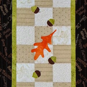 May include: A quilted table runner with a checkered pattern in beige and white. The runner features an orange leaf and brown acorns, with a black and green border with the text "Happy Harvest Giving Thanks" repeated.