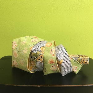 Peut inclure: Quatre paniers de rangement en tissu empilés les uns sur les autres. Les paniers sont en tissu vert à motif floral et bordure jaune. Le panier du haut est plié pour montrer la bordure jaune.