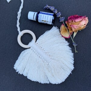 Może przedstawiać: Biały, makramowy odświeżacz powietrza do samochodu w kształcie pióra z drewnianym pierścieniem i małą butelką olejku eterycznego. Butelka jest oznaczona napisem "FRAGRANCE" i ma wzór lawendy i róży.