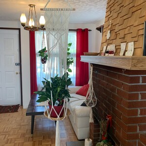 Peut inclure: Un porte-plante en macramé blanc avec une baguette en bois, suspendu au plafond d'un salon. Une plante verte est dans un pot brun à l'intérieur du porte-plante. Il y a un canapé blanc et une cheminée en briques en arrière-plan.