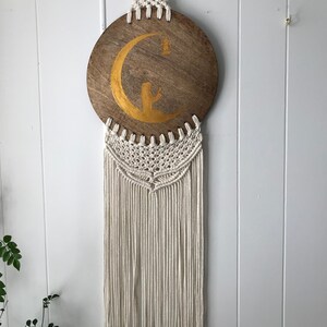 Peut inclure: Une suspension murale en macramé blanc avec un disque rond en bois orné d'un croissant de lune doré et d'une silhouette d'une personne priant.
