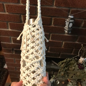 May include: A white crocheted bottle holder with a long handle. The holder is made of thick cotton rope and has a diamond pattern.
