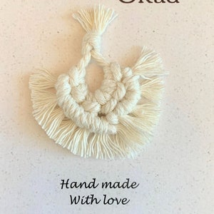 May include: A white macrame wall hanging with a fringe border. The hanging is in the shape of a heart with two circles inside. The text "L'atelier Okad" and "Hand made with love" is visible below the hanging.