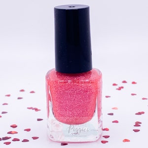 May include: A bottle of pink nail polish with a black cap. The polish has a glittery finish and is surrounded by heart-shaped confetti. The bottle has a clear base with the text "Pure Piggies Polish".