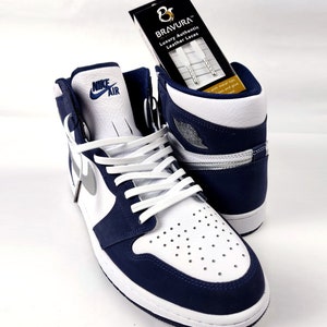 May include: White and navy blue Nike Air Jordan 1 high-top sneakers with white leather laces. The sneakers are on a white surface. A box of white leather laces with the brand name "BRAVURA" is in the background.