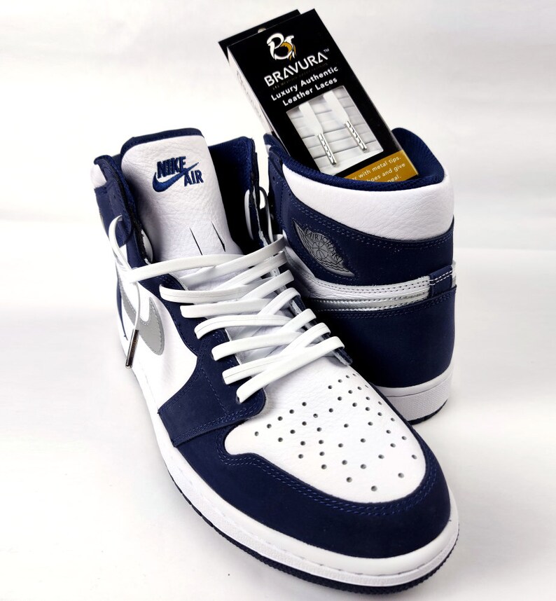 May include: White and navy blue Nike Air Jordan 1 high-top sneakers with white leather laces. The sneakers are on a white surface. A box of white leather laces with the brand name "BRAVURA" is in the background.