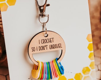 May include: A circular wooden keychain featuring the engraved text I CROCHET SO I DON'T UNRAVEL, attached to a silver metal lobster clasp. A collection of small, colorful plastic stitch markers in yellow, green, pink, and blue hangs from the ring.