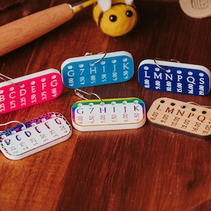May include: A collection of colorful stitch markers with measurements and letters, including pink, blue, and wooden options. Each marker has a safety pin for attachment. A wooden crochet hook and a felt bee are also visible.