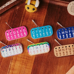 May include: A collection of colorful stitch markers with measurements in inches. Each marker is a small, rectangular tag with a metal loop and needle. The tags are pink, blue, blue, leopard print, rainbow, and tan, with letters and numbers.