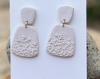May include: A pair of light pink earrings with a snowflake design. The earrings are made of clay and have a rectangular shape with a smaller rectangular top. The earrings are attached to a white card with the text "Skies of Blue studio".