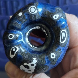 May include: A blue and black donut-shaped ceramic pendant with white swirls and spots. The pendant has a hole in the center.