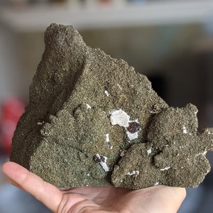 May include: A rough, green and brown rock with white and black mineral inclusions. The rock has a textured surface and appears to be a natural specimen.