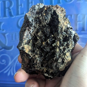 May include: A dark, textured rock formation with a rough, uneven surface. The rock has a mix of dark brown, black, and hints of gold and tan colors. The overall shape is irregular, with a rugged appearance, suggesting a natural mineral specimen.