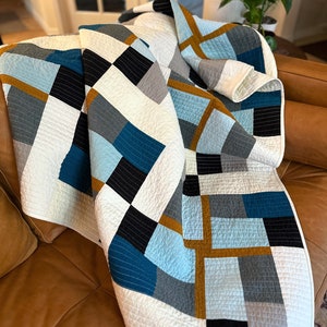 May include: A white, blue, grey, and gold geometric quilt with a modern design. The quilt is folded and draped over a brown leather sofa.