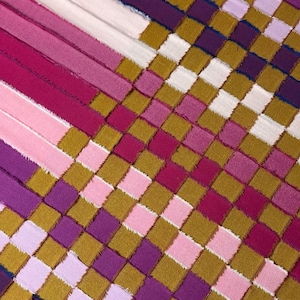 Puede incluir: Una tela a cuadros con cuadrados en tonos de rosa, morado y dorado. La tela está hecha de tiras de diferentes colores cosidas juntas.