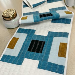 Peut inclure: Un chemin de table et un set de table matelassés avec un motif géométrique. Le motif présente des carrés blancs, bleu sarcelle, noirs et jaune moutarde. Le chemin de table et le set de table sont sur une surface en bois clair.