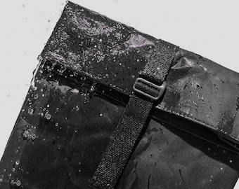 May include: A black waterproof bag with a flap closure and a black adjustable strap. Water droplets are visible on the surface of the bag.