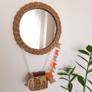 Op de afbeelding: Een ronde rieten spiegel met een bruin geweven frame en een heldere reflectie. De spiegel hangt aan een muur en heeft een klein rieten mandje dat eraan hangt met oranje en bruine vlaggetjes van stof.