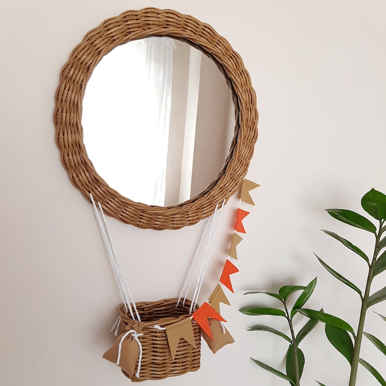 Op de afbeelding: Een ronde rieten spiegel met een bruin geweven frame en een heldere reflectie. De spiegel hangt aan een muur en heeft een klein rieten mandje dat eraan hangt met oranje en bruine vlaggetjes van stof.