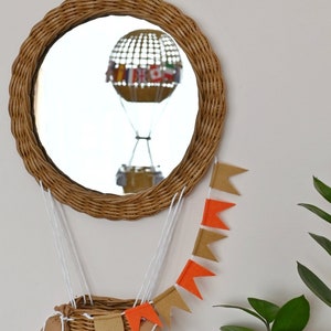 Op de afbeelding: Een rieten spiegel in de vorm van een luchtballon met een witte touwgordijn van oranje en bruine vlaggetjes die eraan hangen. Een rieten mand hangt aan de onderkant van de spiegel.