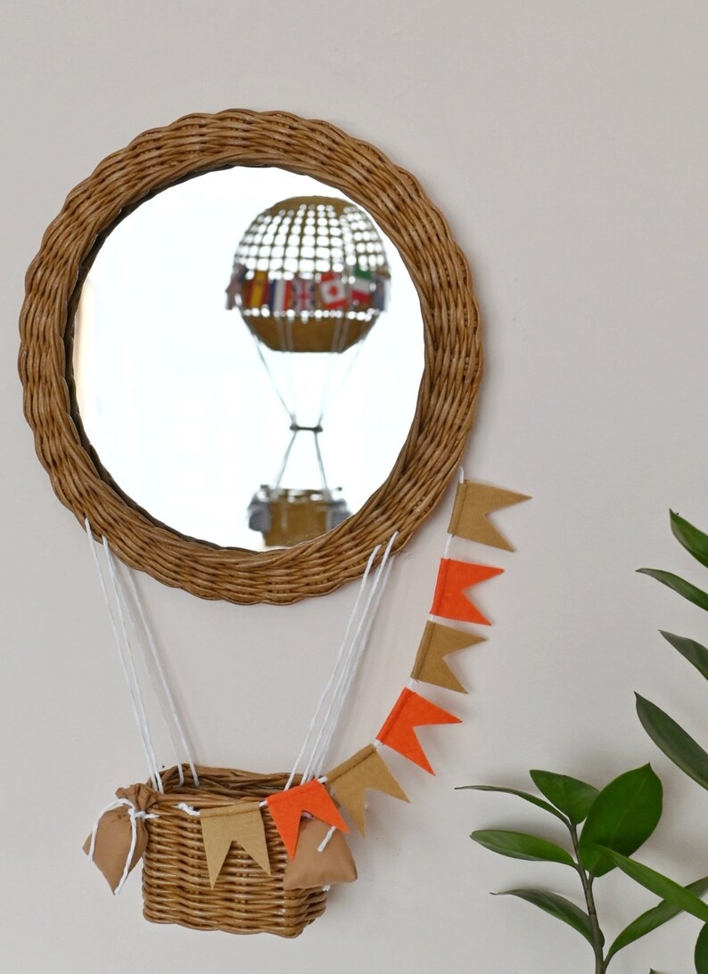 Op de afbeelding: Een rieten spiegel in de vorm van een luchtballon met een witte touwgordijn van oranje en bruine vlaggetjes die eraan hangen. Een rieten mand hangt aan de onderkant van de spiegel.