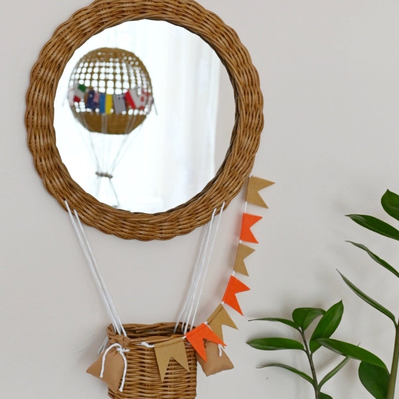 Op de afbeelding: Een ronde spiegel van wilgentenen met een luchtballon die in het glas wordt gereflecteerd. De spiegel hangt aan een witte muur met een slinger van oranje en bruine vlaggen die eronder hangen.