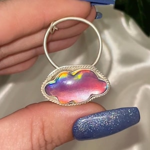 May include: A silver hoop earring with a pink, iridescent cloud charm. The cloud is set in a silver bezel and has a textured edge.