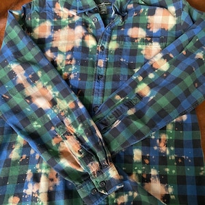 May include: A blue and green plaid flannel shirt with bleach splatters. The shirt has a button-up front and long sleeves.