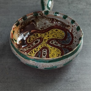 Puede incluir: Un cuenco de cerámica blanca con un asa verde y marrón. El cuenco tiene un diseño abstracto colorido en tonos de amarillo, marrón y azul.