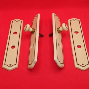 Op de afbeelding: Set van vier antieke porseleinen deurknoppen met zwarte rand en witte keramische knoppen. De knoppen hebben de vorm van een rechthoek met afgeronde hoeken.