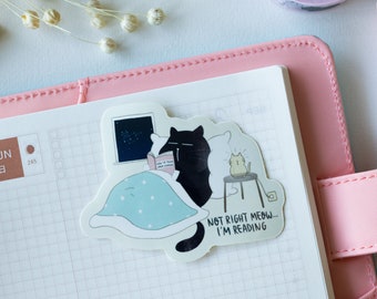 May include: A sticker showing a black cat reading in bed, with the text "NOT RIGHT MEOW... I'M READING." The sticker is on a pink planner with a grid-lined page. A small cat is on a stool next to the bed.