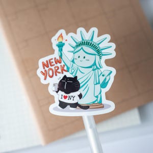 Pode incluir: Um autocolante com a Est&aacute;tua da Liberdade e um gato preto a tirar uma selfie. A Est&aacute;tua da Liberdade &eacute; azul clara, segurando uma tocha e um tablet. O gato usa uma camisa com "I ❤️ NY" e as palavras "NEW YORK" est&atilde;o acima.