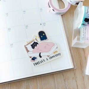Pode incluir: Um autocolante com um gato preto a dormir numa cama com uma manta rosa, uma janela com estrelas e o texto "FOREVER A HOMEBODY". O autocolante est&aacute; num calend&aacute;rio.