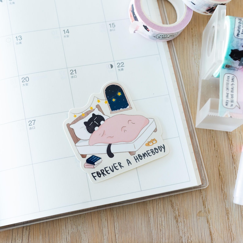 Pode incluir: Um autocolante com um gato preto a dormir numa cama com uma manta rosa, uma janela com estrelas e o texto "FOREVER A HOMEBODY". O autocolante est&aacute; num calend&aacute;rio.