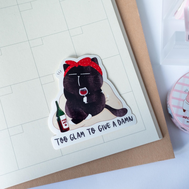 Pode incluir: Um autocolante com um gato preto com uma bandana vermelha, segurando uma ta&ccedil;a de vinho e uma garrafa de vinho. O texto no autocolante diz "TOO GLAM TO GIVE A DAMN" e "WINE NOT". O autocolante est&aacute; num calend&aacute;rio.