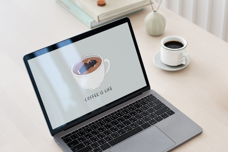 Op de afbeelding: Een laptop met een grijze buitenkant en een zwart toetsenbord. Het scherm toont een cartoonafbeelding van een kopje koffie met een slapende kat erin. De tekst "COFFEE IS LIFE" wordt onder de afbeelding weergegeven.