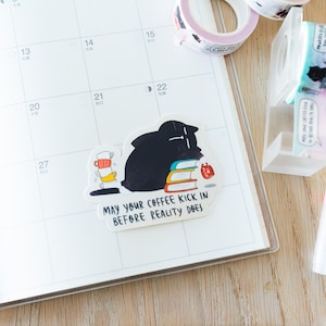 Puede incluir: Una pegatina de un gato negro con una pila de libros y una taza de café. La pegatina tiene el texto "May your coffee kick in before reality does."