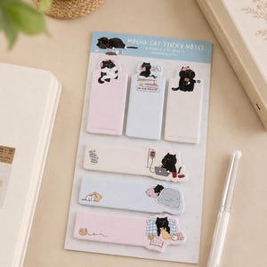 May include: A set of Mocha Cat sticky notes on a backing card featuring six rectangular adhesive strips in pastel pink, blue, and white. Each strip displays a black cat illustration engaged in various activities like reading, sleeping, or working on a laptop.