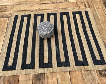 May include: A rectangular area rug featuring a natural tan fiber base with a bold, black geometric maze pattern. Centered on the rug is a small, round, upholstered footstool with a detailed black and white intricate floral print.
